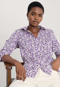 Lila, virágmintás gombos ing fehér és tengerészkék színekkel. Hosszú ujjai felgöngyölve. Simább anyagtextúra. Világos színű nadrággal viselve.