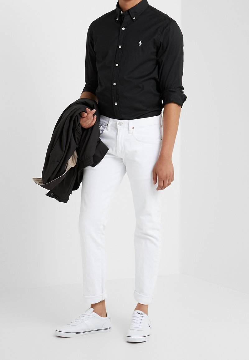 Czarna koszula na guziki, białe dżinsy z denimu, białe trampki. Osoba trzyma czarną kurtkę. Prosty fason, regularny krój, bez widocznych wzorów.