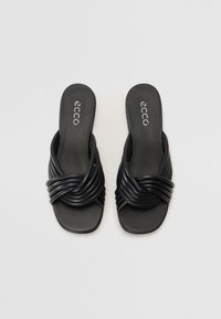 Des sandales à glissière noires en cuir lisse, présentant un design de lanière torsadée et une semelle plate. Le logo est visible sur le pied.