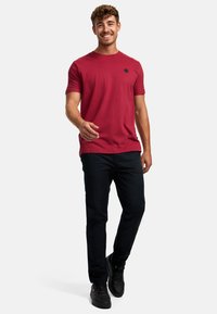 Rød bomulls T-skjorte med rund hals og korte ermer, med et lite svart logodetalj. Brukt med svarte bukser og joggesko.