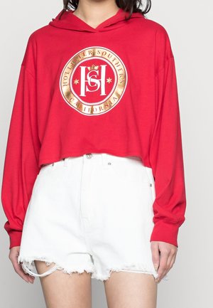 Personne portant un sweat à capuche court rouge avec le logo Hollister Southern California et un short en jean blanc taille haute aux bords effilochés.