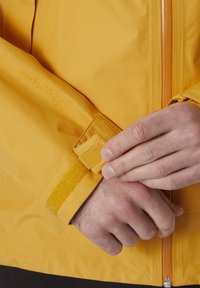 Geel waterdichte jas van een gestructureerde stof, met een klittenband manchetverstelling en een rits aan de voorkant. Biedt een close-upweergave van de details.