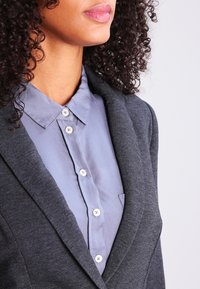 Dark gray blazer over a light blue shirt with a pointed collar and button closures, featuring a smooth texture and subtle stitching details.