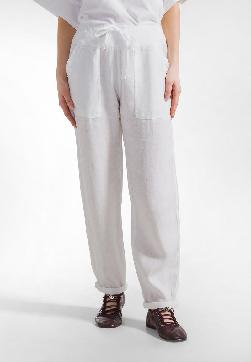 Valkoiset pellavahousut, joissa on kiristysnauha vyötäröllä, kaksi etutaskua ja käännetyt lahkeet. Yhdistetty ruskeisiin nauhallisiin kenkiin.