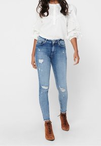 Kvinna i vit långärmad blus med spetsdetaljer, ljusblå slitna skinny jeans och bruna mocka snörstövlar.