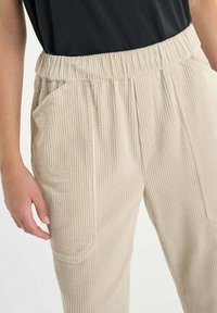 Beige corduroy broek met een elastische tailleband, voorzien van zijzakken en een geribbelde textuur. Gefocuste weergave van de taille naar beneden.