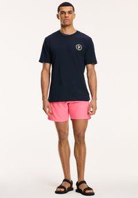 Tengerészkék, rövid ujjú póló, kör alakú logóval, élénk rózsaszín fürdőshorts-szal és fekete szandállal párosítva. A modell egy semleges háttérben áll.
