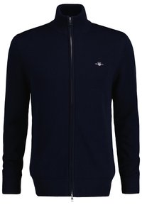 Pull-over zippé marine avec un col montant, détails côtelés aux poignets et à l'ourlet, et un petit emblème sur la poitrine gauche. Texture en tricot douce.