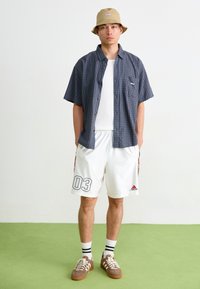 Chemise à manches courtes à carreaux bleus et noirs, débardeur blanc, shorts blancs avec des accents rouges et noirs, baskets marron et chapeau de paille beige.