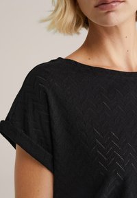 Černý strukturální top s herringbonovým vzorem, krátkými ohrnutými rukávy a širokým výstřihem. Vyrobeno z lehké, prodyšné tkaniny.