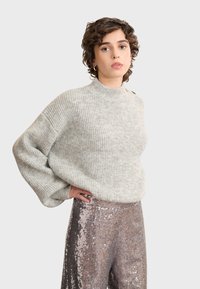 Svijetlosiva rebrasta pletena majica s visokim izrezom i oversized rukavima, u kombinaciji s hlačama ukrašenim sjajnim sequins detaljima.