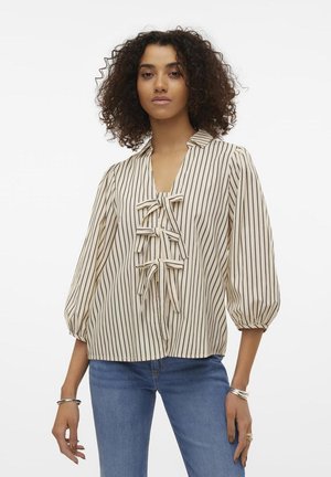 Ljusbeige blus med vertikala svarta ränder, med V-ringning, puffärmar och en mittdetalj i form av en rosett. Buren över blå jeans.