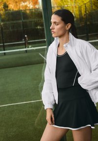 Bílá zipová bunda s balónovými rukávy přes černé sportovní šaty s rozšířenou sukní a bílým lemováním. Na pozadí tenisového kurtu.