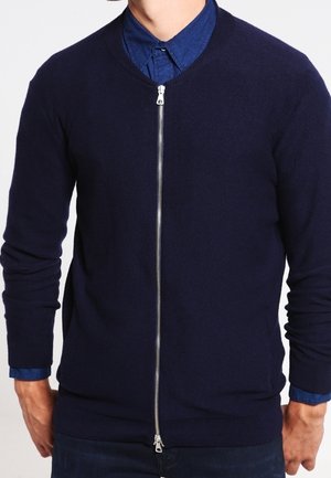 Mężczyzna w granatowym kardiganie na zamek, noszący pod spodem ciemnoniebieską koszulę zapinaną na guziki, z rękami swobodnie opuszczonymi wzdłuż ciała.