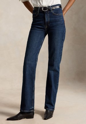 Modèle portant un jean bleu foncé à jambes droites, une ceinture noire, une chemise blanche et des bottines noires à bout pointu.