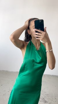 Vestido halter verde com um design drapeado, textura suave e leve brilho. Modelo segura um smartphone, com paredes e chão brancos visíveis.
