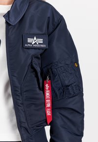 Mørkeblå bomberjakke med glidelås, ribbestrikkede mansjetter og et brodert logo-patch. Har en verktøylomme og et rødt merke med teksten "FJERN FØR FLYGING."