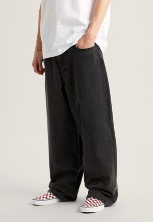 Person trägt lockere schwarze Hose, weißes T-Shirt und rot-weiß karierte Slip-On-Schuhe, mit einer Hand in der Tasche.