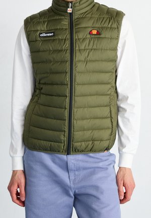 Homme portant une veste sans manches matelassée vert olive avec fermeture éclair et écussons du logo de la marque par-dessus une chemise blanche à manches longues et un pantalon bleu clair.