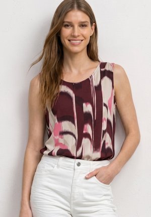 Femme souriante aux longs cheveux portant un haut sans manches à motif abstrait violet et rose, rentré dans un pantalon blanc, main dans la poche, contre un mur blanc.