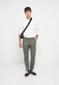 Pantalons cargo verts avec fermeture à bouton, dotés de poches latérales, associés à une chemise blanche à manches longues texturée et des baskets noires.