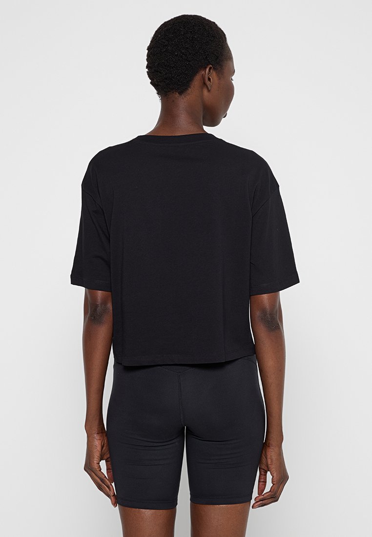 Persona con capelli rasati indossa una camicia nera a maniche corte larga e pantaloni da ciclismo neri attillati, vista da dietro su uno sfondo neutro.