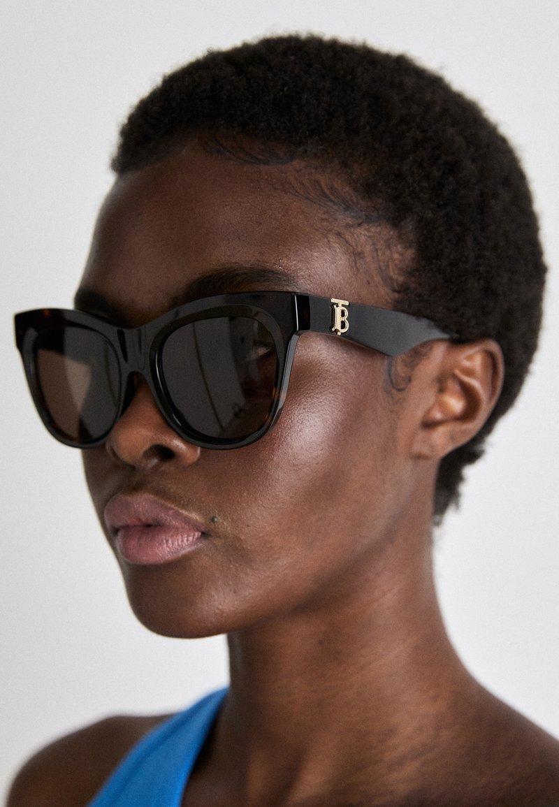 Lunettes de soleil noires avec une monture épaisse et angulaire, dotées d'un logo doré "TB" sur le côté et de verres teintés sombres, sur un fond neutre.