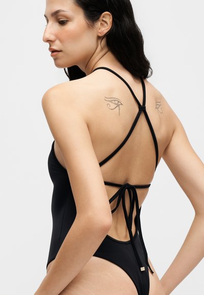 Femme aux longs cheveux foncés portant un maillot de bain noir à bretelles, montrant son dos avec des tatouages d'yeux symétriques sur les deux omoplates.