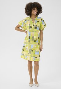 Īsām piedurknēm tērps dzeltenā krāsā ar ģeometrisku rakstu, kurā redzami kvadrāti zaļā, zilā un baltā krāsā. Brīvi pieguļošs, bez pogām ap kaklu.