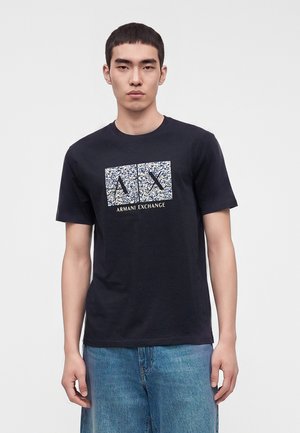 Jeune homme portant un t-shirt noir Armani Exchange avec un logo AX bleu et blanc tacheté et un jean bleu clair, debout devant un fond uni.