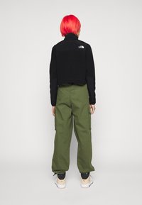 Giacca in pile nera cropped con colletto alto, pantaloni cargo verde oliva e sneakers beige. Presenta un dettaglio del logo sulla giacca.