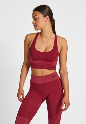Femme aux cheveux tressés portant un soutien-gorge de sport rouge assorti et un legging de la marque Hummel, debout et regardant sur le côté sur un fond uni.