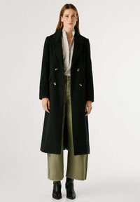 Cappotto doppiopetto in lana nera con colletto a lancia, bottoni argentati e design lungo, abbinato a pantaloni larghi verde chiaro e stivaletti.