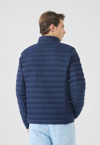 Hombre vestido con una chaqueta acolchada azul marino y jeans de un azul claro, de espaldas contra un fondo liso claro.