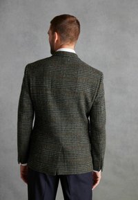 Tamsiai žalia tweed švarkas su rudos ir mėlynos spalvos čekio raštu, struktūruota siluetas, iškirptės apykaklės ir šoninės atramos, vilkimas virš tamsių kelnių.