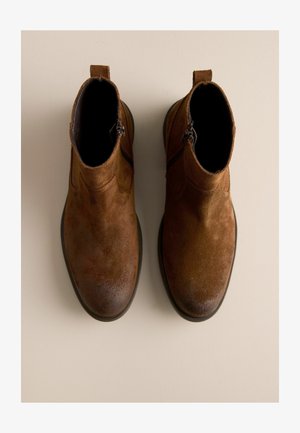 Braune Wildleder-Stiefeletten mit glatter Textur, seitlichen Reißverschlüssen, runder Zehenpartie und robuster schwarzer Sohle. Klassisches Design mit minimalen Akzenten.