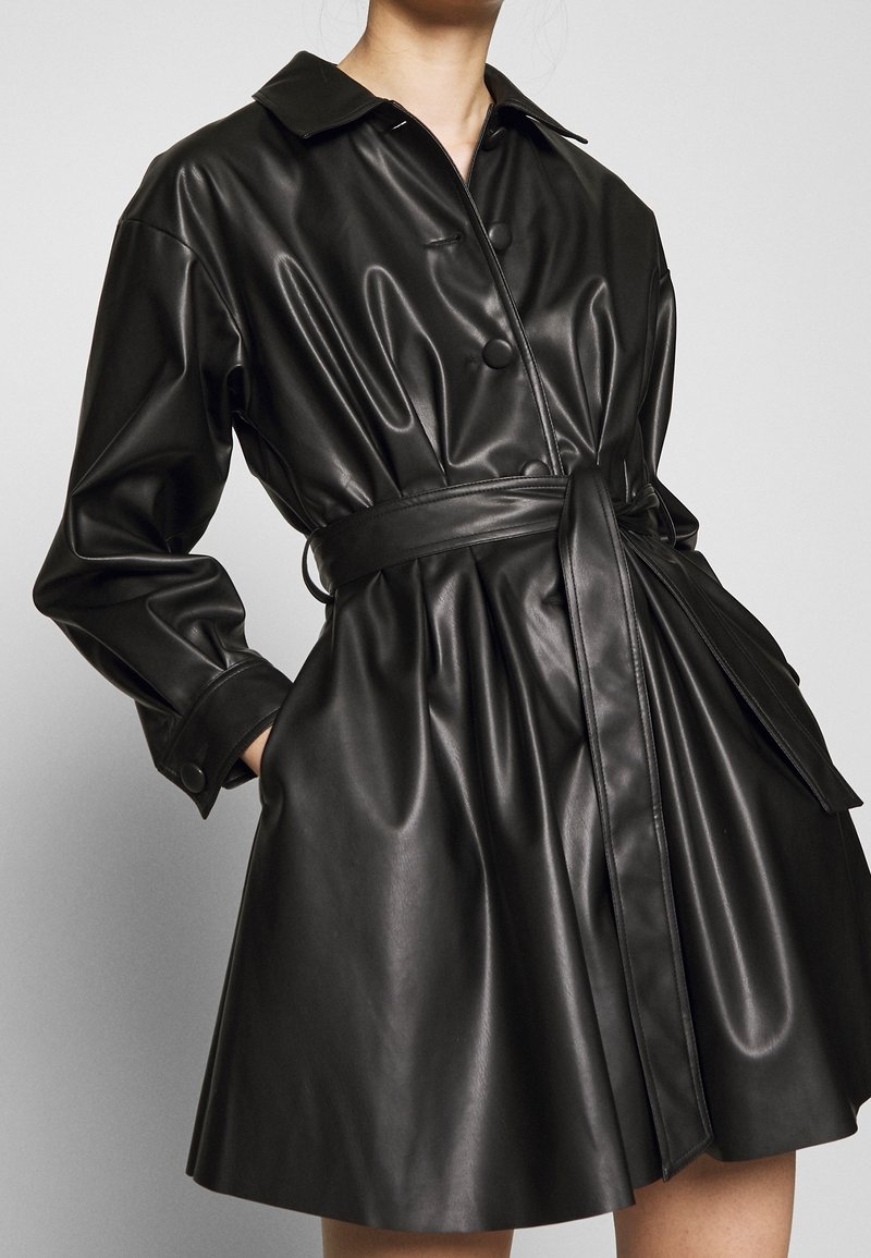 Kobieta w czarnej sukience z imitacji skóry z długimi rękawami, zapięciem na guziki, kołnierzykiem i dopasowanym paskiem wiązanym w talii.