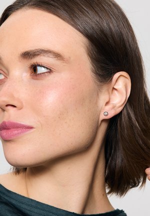 Okrągły, srebrny kolczyk wkręcany z przejrzystym kamieniem szlachetnym. Zbliżenie ucha modelki, pokazujące gładką strukturę skóry i delikatne piegi.