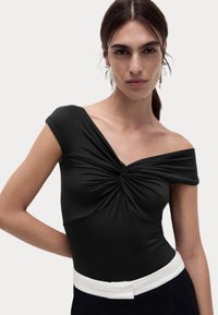 Sort off-the-shoulder top lavet af glat stof med en snoet detalje ved brystet og et hvidt taljebånd, der skaber en tætsiddende silhuet.