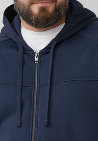 Marineblaues Zip-Hoodie aus strukturiertem Material, mit einer doppellagigen Kapuze, Zugbändern und silbernen Reißverschlussdetails auf der Vorderseite.