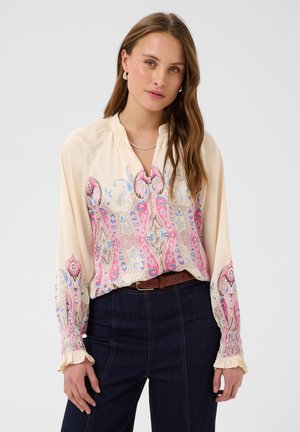 Femme aux longs cheveux bruns portant un chemisier crème à motifs cachemire roses, un jean foncé, une ceinture marron et des boucles d’oreilles en perles, debout face à l’avant.