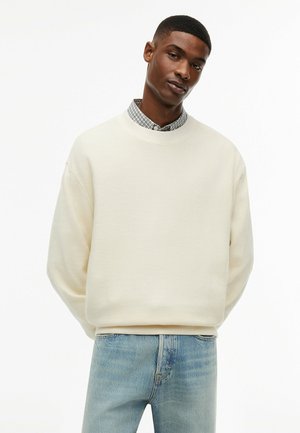 Mann trägt einen cremefarbenen Strickpullover über einem karierten Hemd, kombiniert mit hellblauen Jeans, steht mit den Händen hinter dem Rücken.
