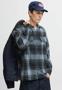 Camisa de franela a cuadros en tonos gris y negro, con dos bolsillos en el pecho, botones al frente y mangas largas, combinada con una chaqueta de mezclilla.