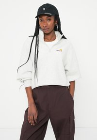 Felpa grigia chiara con zip a quarti e strato interno bianco, con accenti del logo arancione. Abbinata a pantaloni cargo marrone scuro e un cappellino nero.