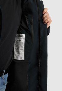 Jachetă neagră cu un exterior din textile neted, având un buzunar cu fermoar și o etichetă vizibilă în interior. Interiorul prezintă o culoare contrastantă.
