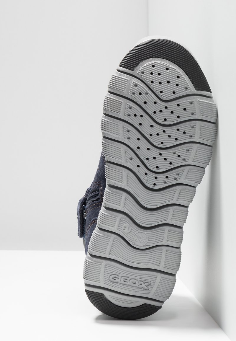 Granatowe buty z tkaniny z gumową podeszwą, z szaro teksturowaną podeszwą zewnętrzną i żłobionymi wzorami. Rzep zapewniający pewne dopasowanie.