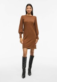 Robe en tricot marron avec un col rond et des manches longues bouffantes, assortie à des bottes noires à talons hautes jusqu'au genou. Texture lisse, design longueur genoux.