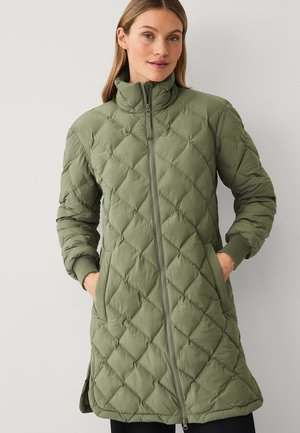 Vrouw draagt een olijfgroene doorgestikte jas tot op de knieën met een ritssluiting aan de voorkant en steekzakken, staand tegen een effen lichte achtergrond.