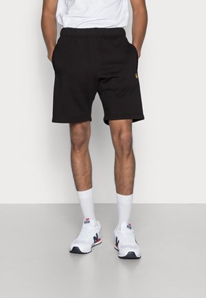 Mand iført sorte shorts, hvide sokker og hvide New Balance-sneakers, stående med hænderne i lommerne mod en ensfarvet baggrund.