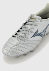 Botină de fotbal din piele gri deschis cu finisaj texturat, având accente cu dungi negre, design cu șireturi și cuie modelate.